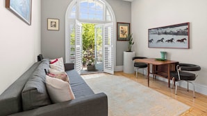 A modern living room with a grey sofa, a wooden dining table with chairs, and a large window with shutters. - Duffotel Paddington LUXE (Paddington)