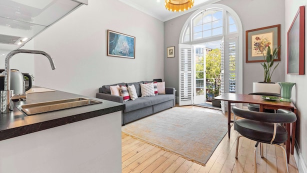 A modern kitchen with a built-in sink, a dining area with a table and chairs, a gray sofa with cushions, and a large window with shutters. - Duffotel Paddington LUXE (Paddington)