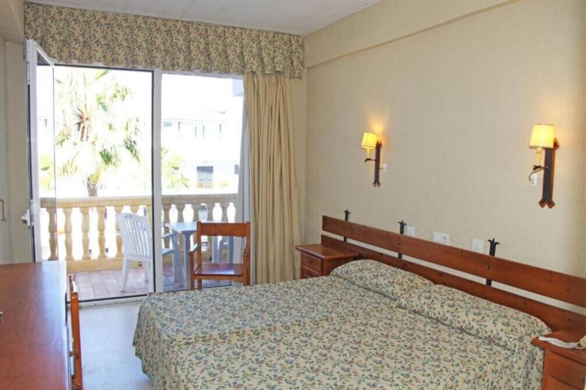 A bedroom with a bed, a desk, a chair, and a balcony with a view of palm trees.