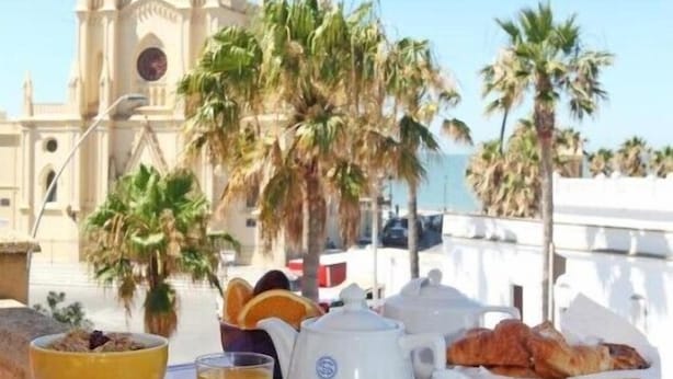 Une table dressée pour le petit-déjeuner, avec un panier de croissants, un bol de nourriture et une théière, le tout sur fond d’une église et de palmiers.