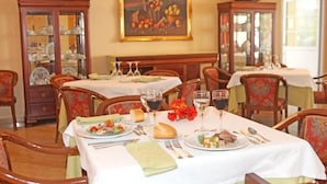 Une salle à manger avec plusieurs tables dressées pour les repas, chacune étant agrémentée d’assiettes, de couverts et de verres. On y trouve des chaises en bois et, au fond, un buffet dont les portes sont en verre.