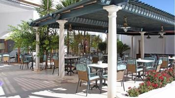 Un patio al aire libre con mesas y sillas, rodeado de palmeras y una pérgola cubierta.
