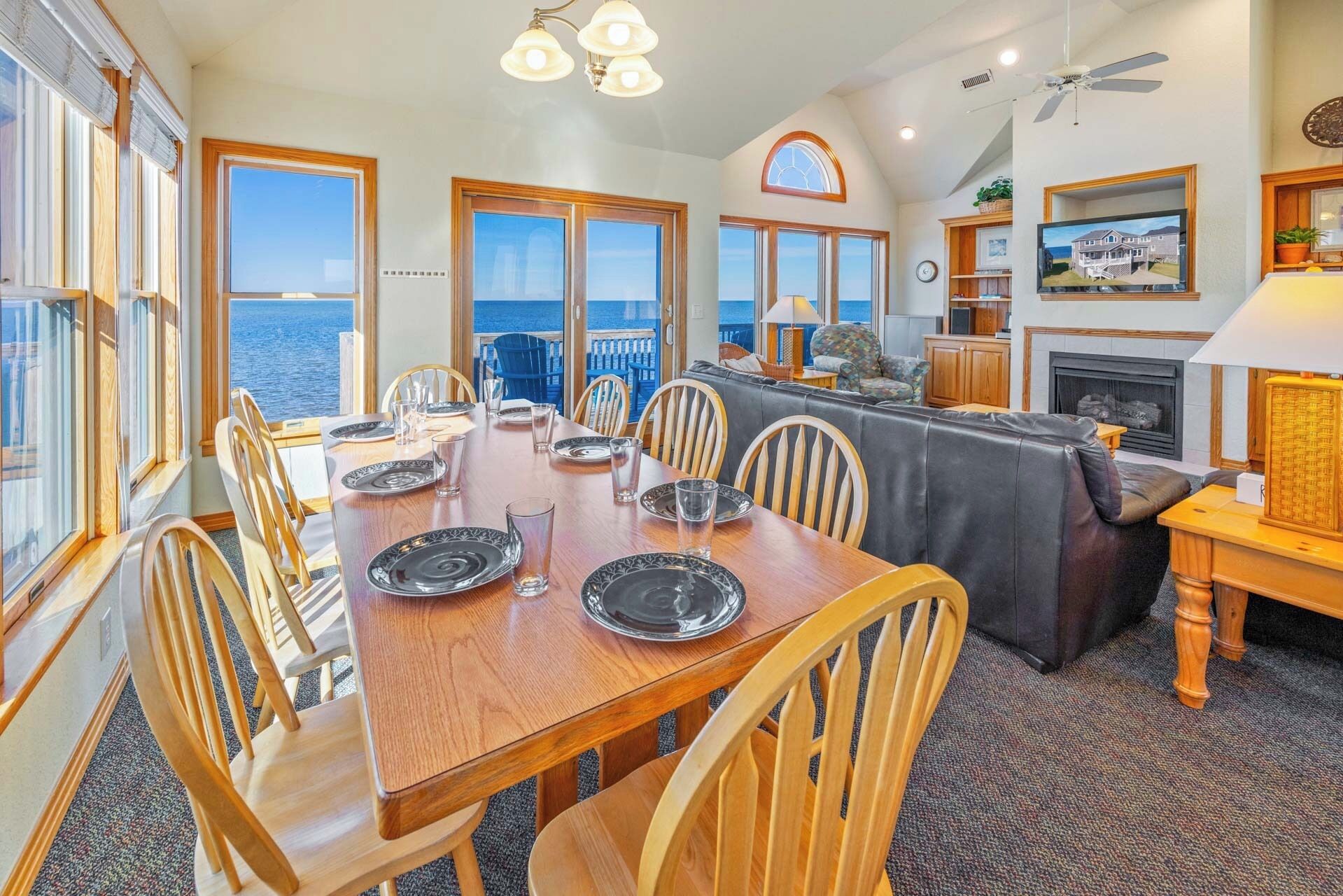 Comedor con mesa de madera dispuesta para seis personas, ventanas amplias con vistas al océano, chimenea y ventilador de techo.