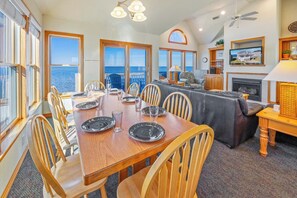 A dining area with a wooden table set for six, large windows with ocean views, a fireplace, and a ceiling fan. - Soundfront w/ Hot Tub, Media Room, Windsports Access, Community Pool/Boat Dock (Avon)