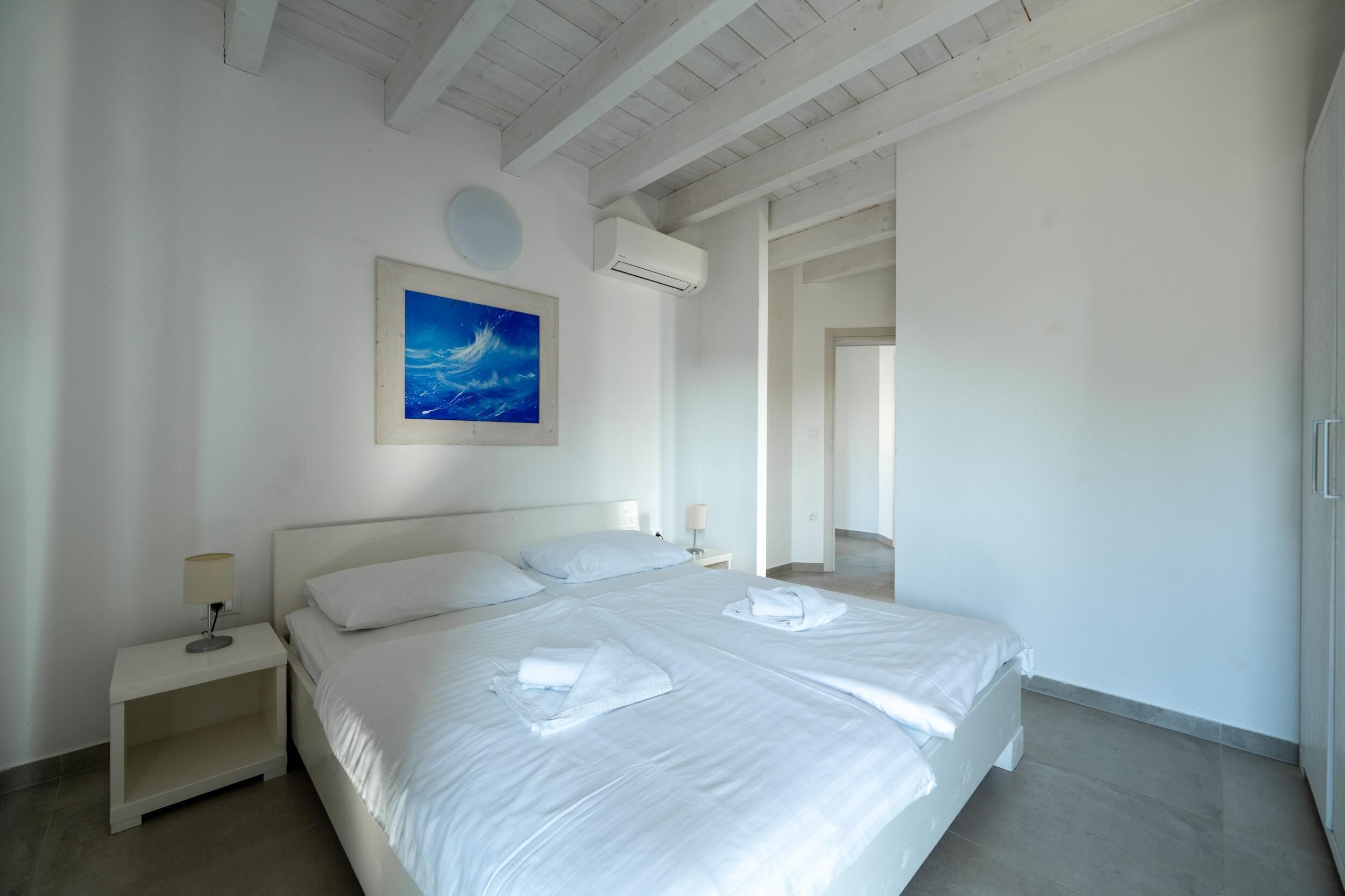 Un grand lit, bien fait, avec des draps blancs et deux oreillers, une table de chevet avec une lampe, et un tableau représentant un ciel bleu avec des nuages accroché au mur.