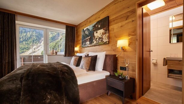 A bedroom with a large bed, wooden walls, a painting, and a view of a mountainous landscape. - Residenz am Lech - Wohnung 3 (Steeg, Lechtal)