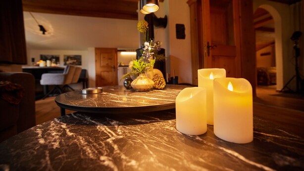 A dining table with candles and a vase of flowers in a warmly lit room with wooden ceilings and a kitchen in the background. - Residenz am Lech - Wohnung 4 (Steeg, Lechtal)
