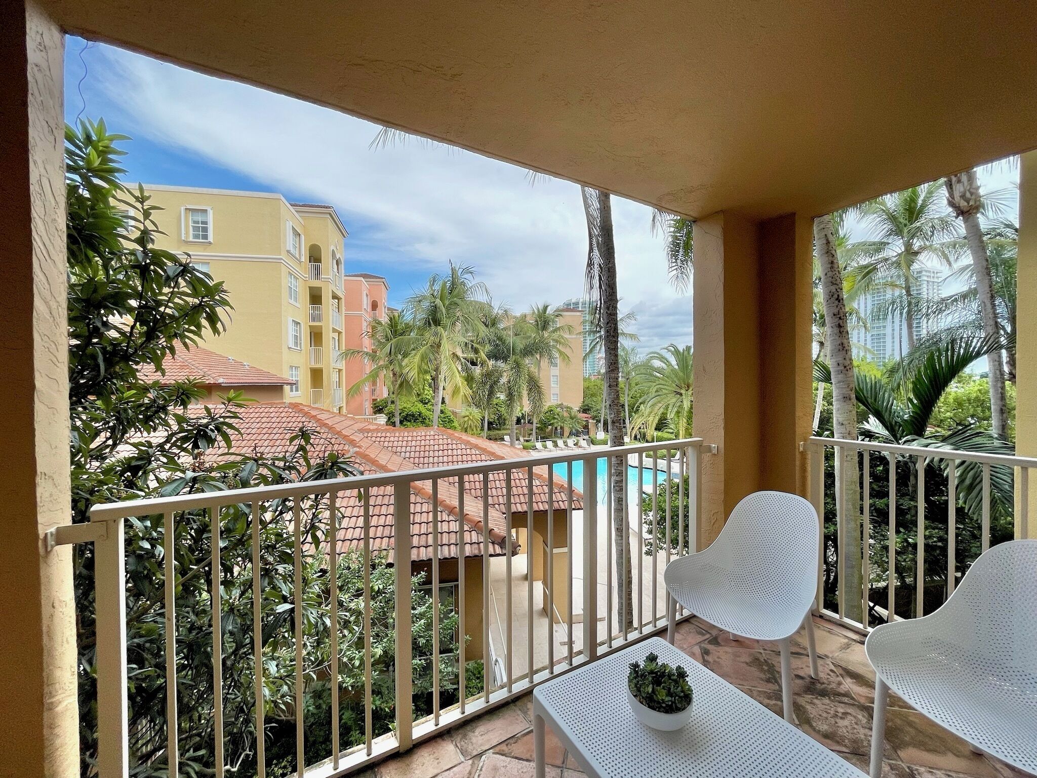 Un balcon avec des balustrades en métal blanc, une table blanche ornée d’un pot de fleurs et des chaises blanches.