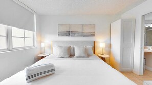 Habitación de hotel con una cama grande, dos mesitas de noche con lámparas, un cabecero, un cuadro sobre la cama y una puerta que da al baño.