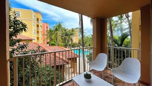 A balcony with two white chairs and a small table, overlooking a swimming pool and buildings with red-tiled roofs.