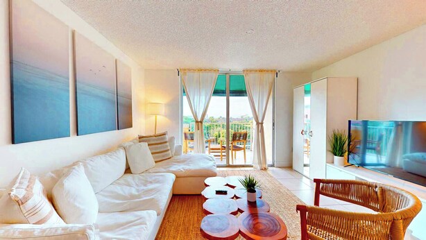 A modern living room with a sofa, coffee table, and TV. There is a balcony with chairs and a view of greenery.