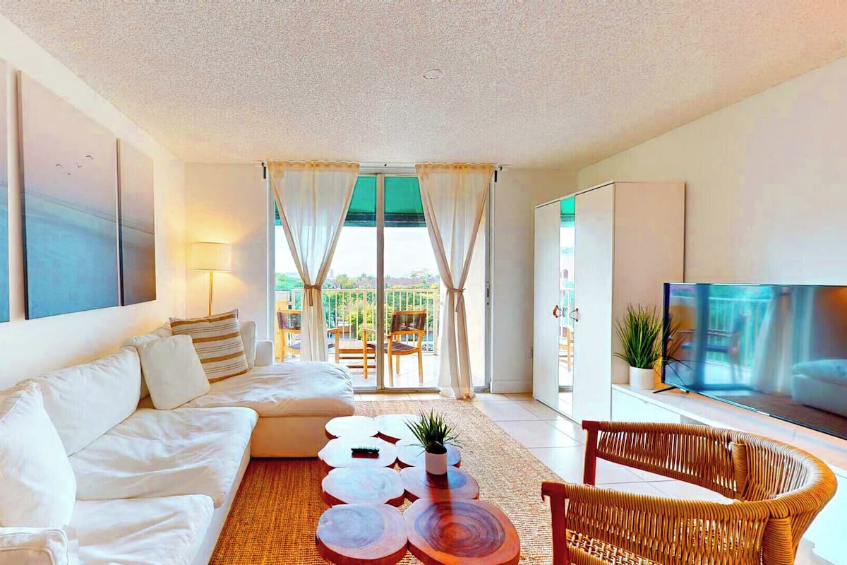 Una sala de estar moderna con sofá, mesa de centro y televisor. Hay un balcón con sillas y vista a zonas verdes.
