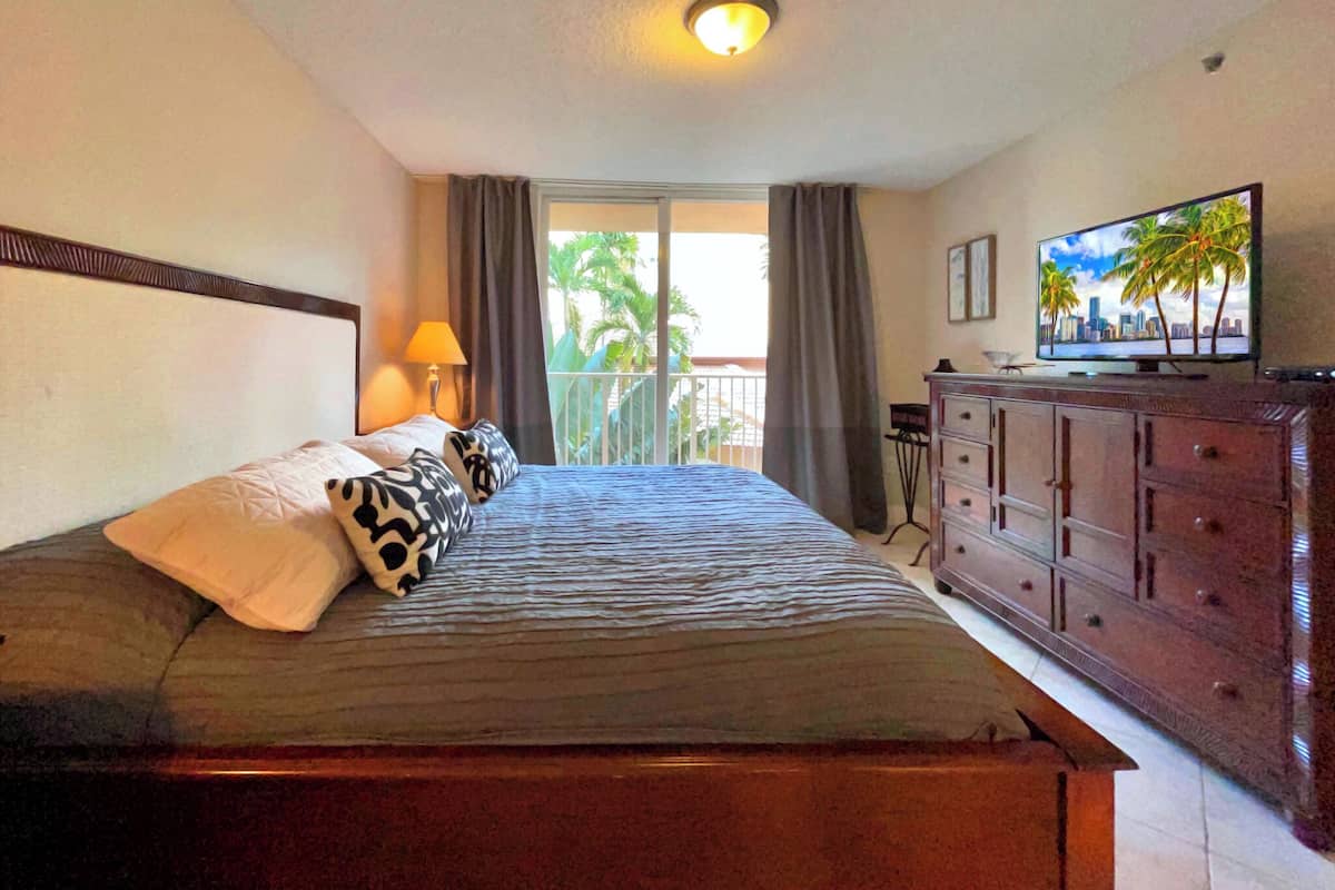 Habitación de hotel con cama, cómoda, televisión y balcón con vistas a palmeras.
