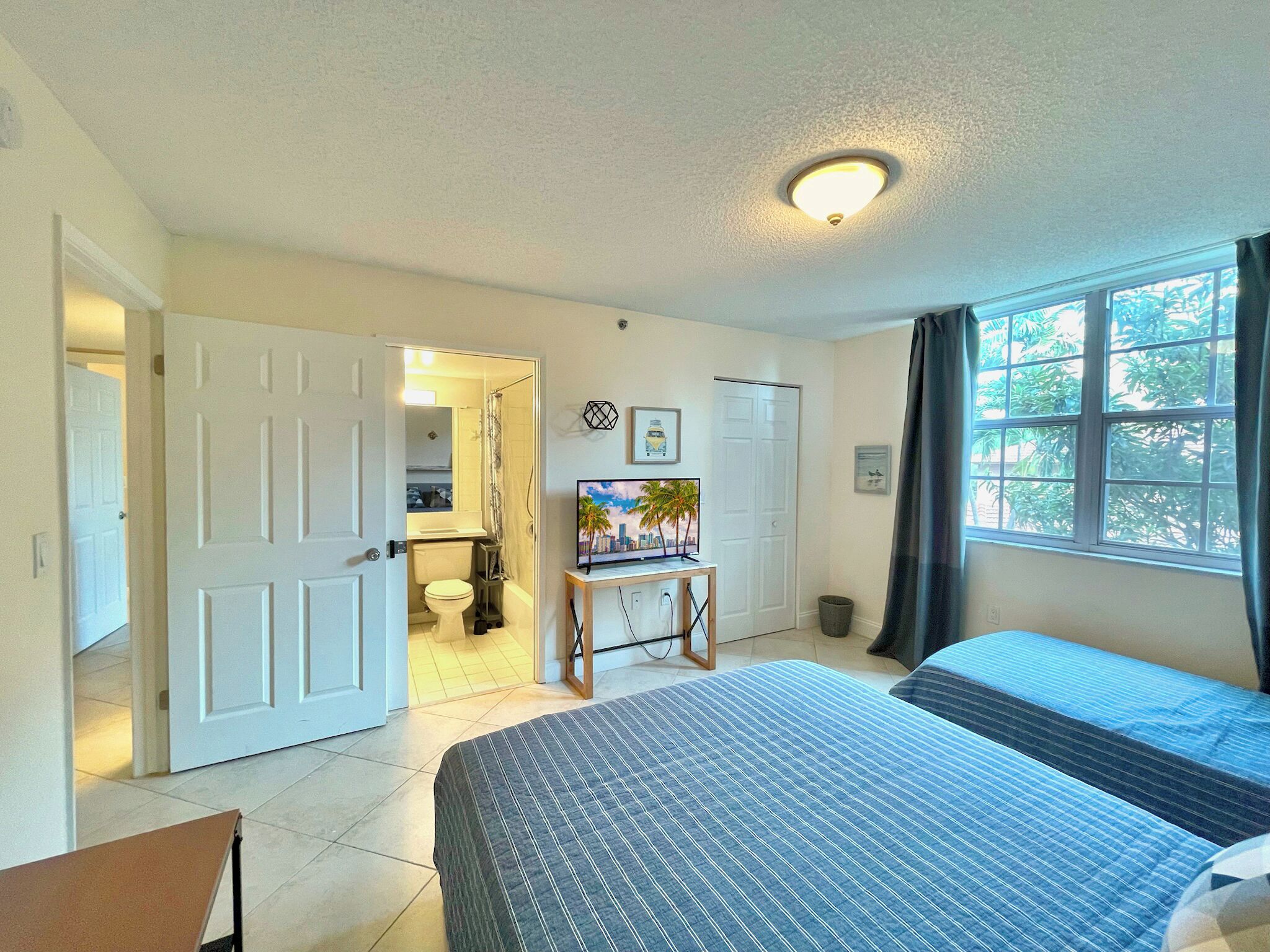 A bedroom with a bed, a TV on a stand, a door leading to a bathroom, and a window with curtains.