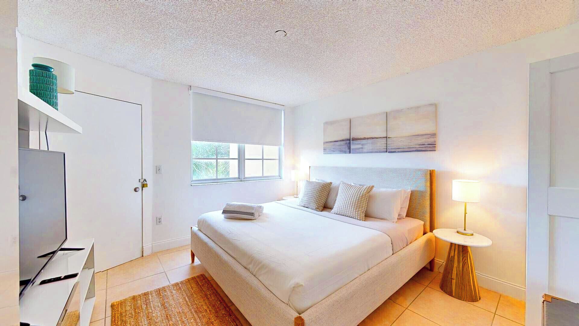 Habitación de hotel con cama, televisor, ventana y un cuadro en la pared.