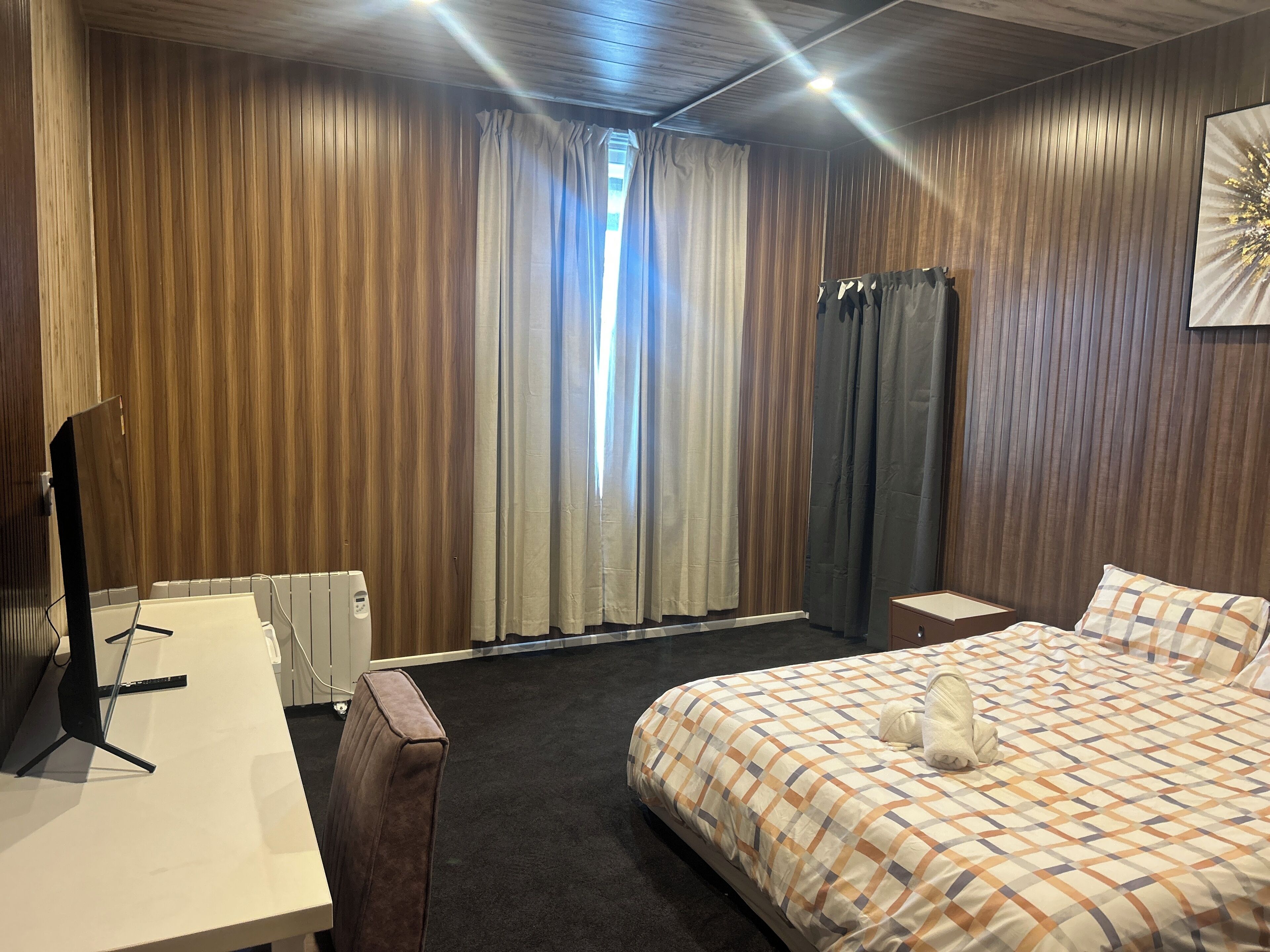 Quarto de hotel com cama, escrivaninha, cadeira, radiador e janela com cortinas.