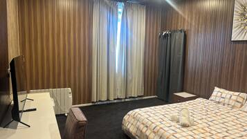 Quarto de hotel com cama, escrivaninha, cadeira, radiador e janela com cortinas.