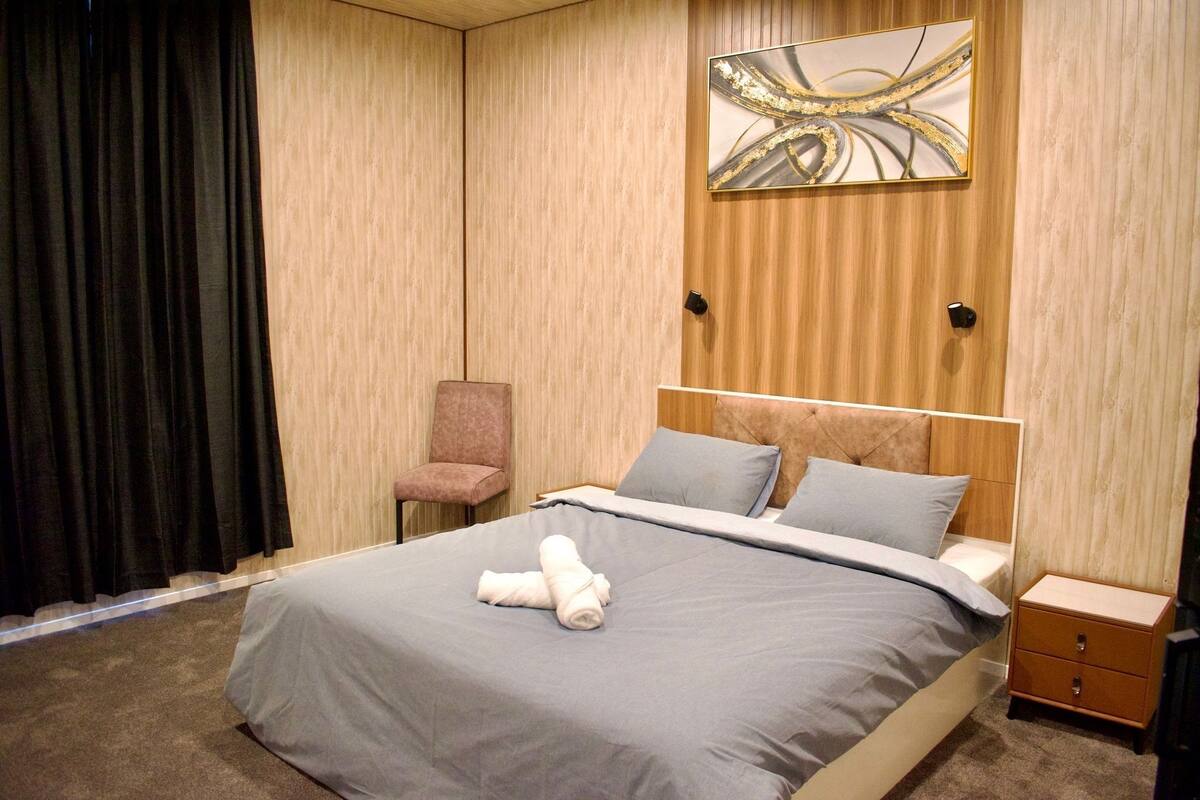 Una cama bien hecha con edredón gris y blanco, un par de toallas enrolladas y un cabecero de madera. A la derecha, una mesita de noche de madera con cajones.