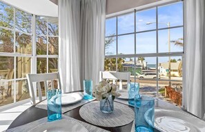 A dining table with blue glassware, white plates, and a vase with white flowers, set against a backdrop of large windows with a view of palm trees and buildings.