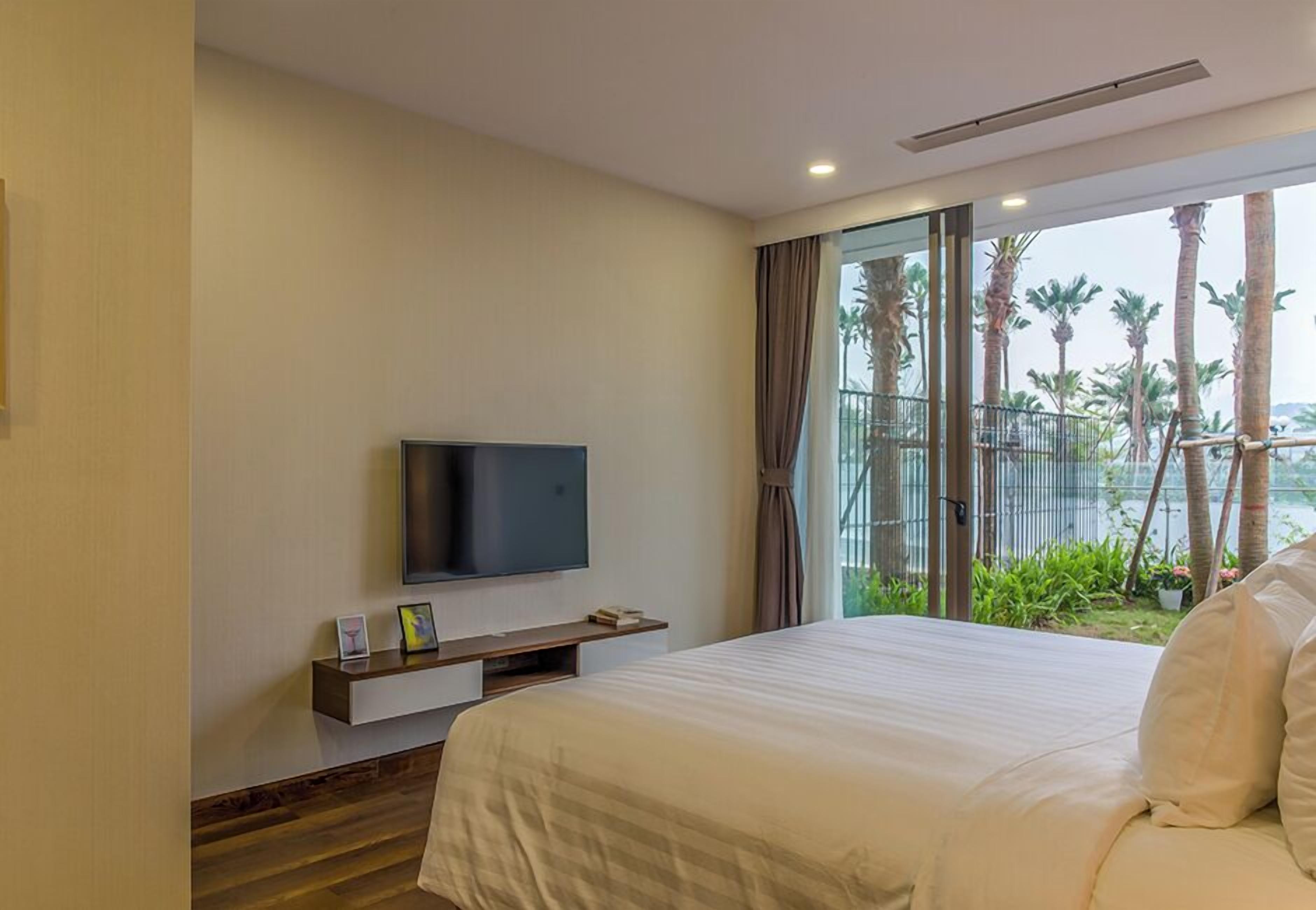 Habitación de hotel con una cama grande, un televisor de pantalla plana sobre un mueble de madera y un balcón con vistas a palmeras.
