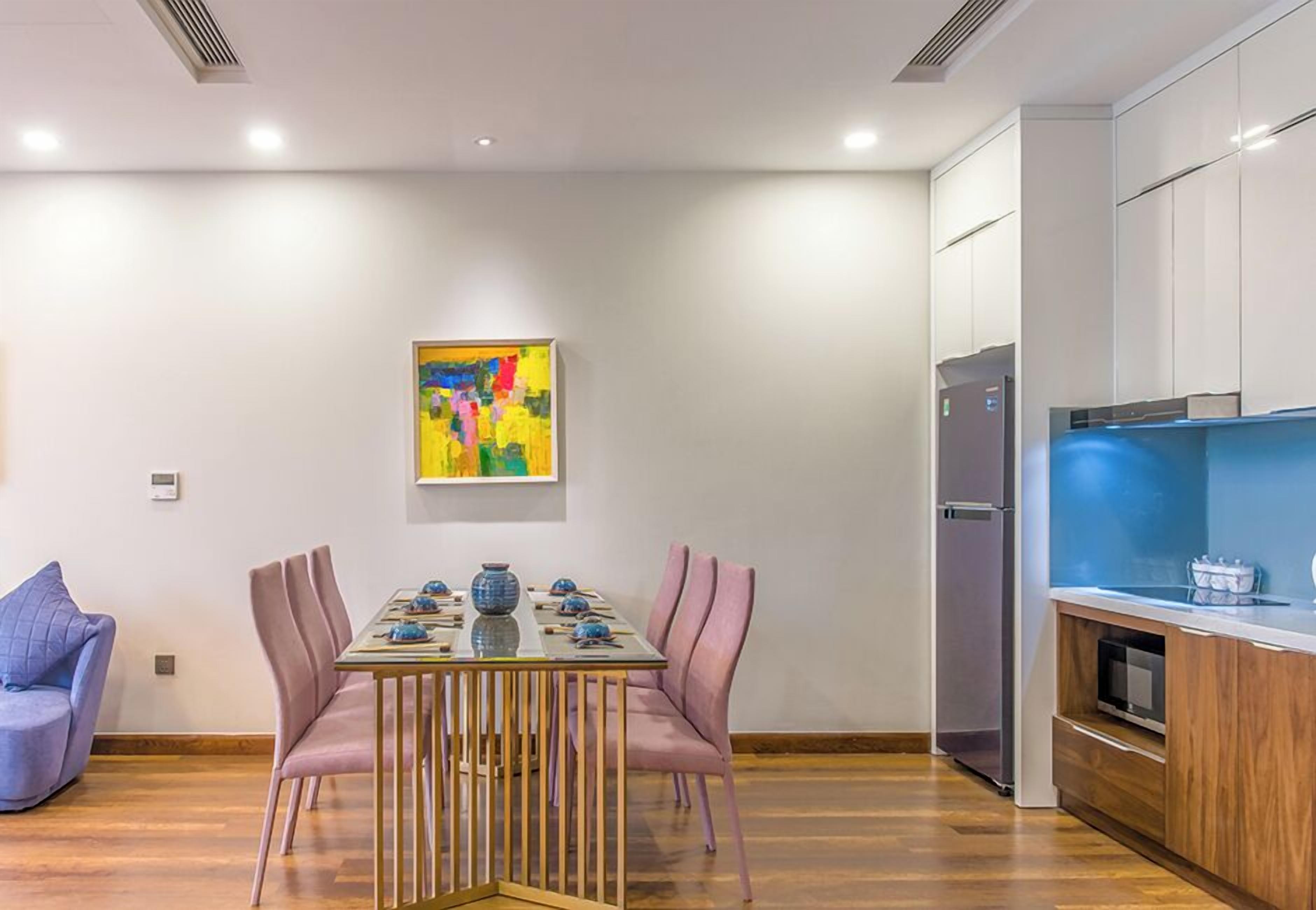 A modern dining area with a wooden table and pink chairs, a colorful abstract painting on the wall, and a kitchen with white cabinets and a blue countertop.