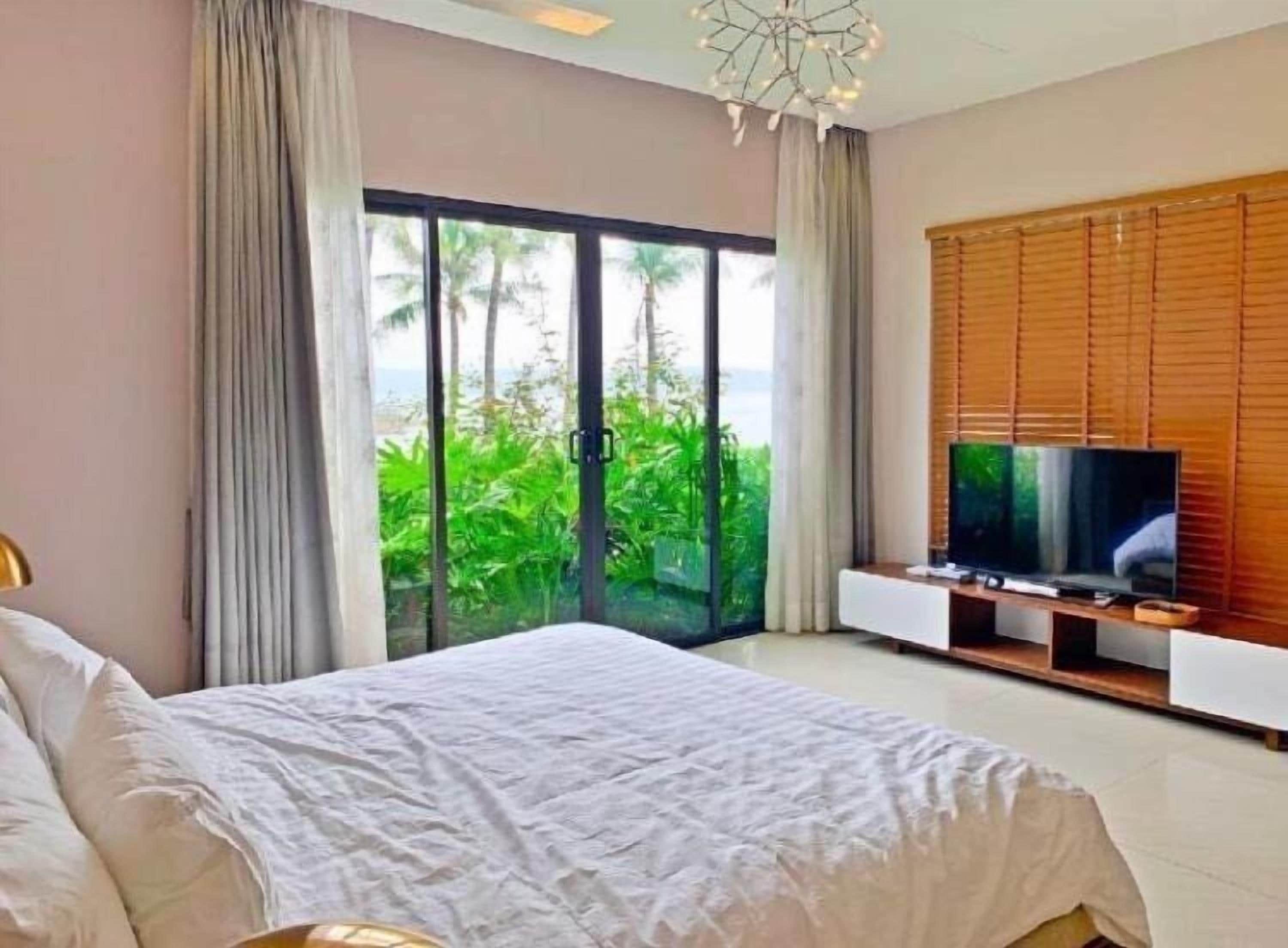 A bedroom with a bed, a TV on a wooden stand, and a sliding glass door leading to a balcony with greenery.