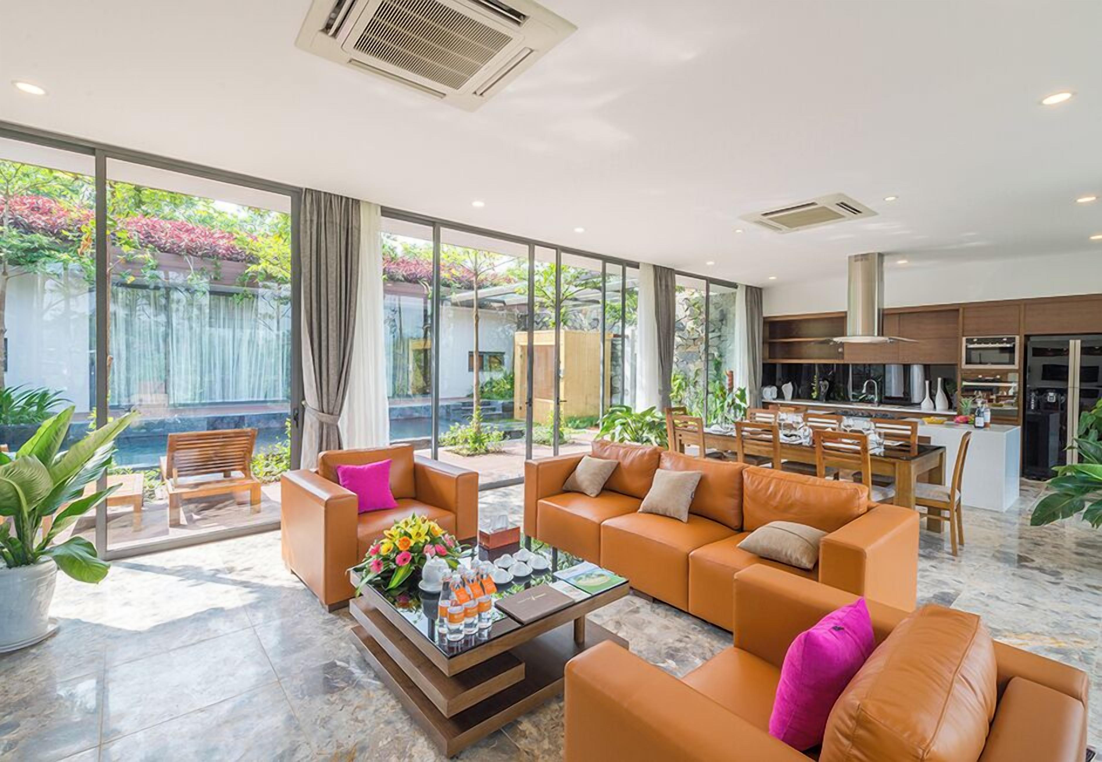 Un salon moderne avec des canapés orange, une table basse avec des boissons, et une vue sur une cuisine et un espace extérieur agrémenté de plantes.