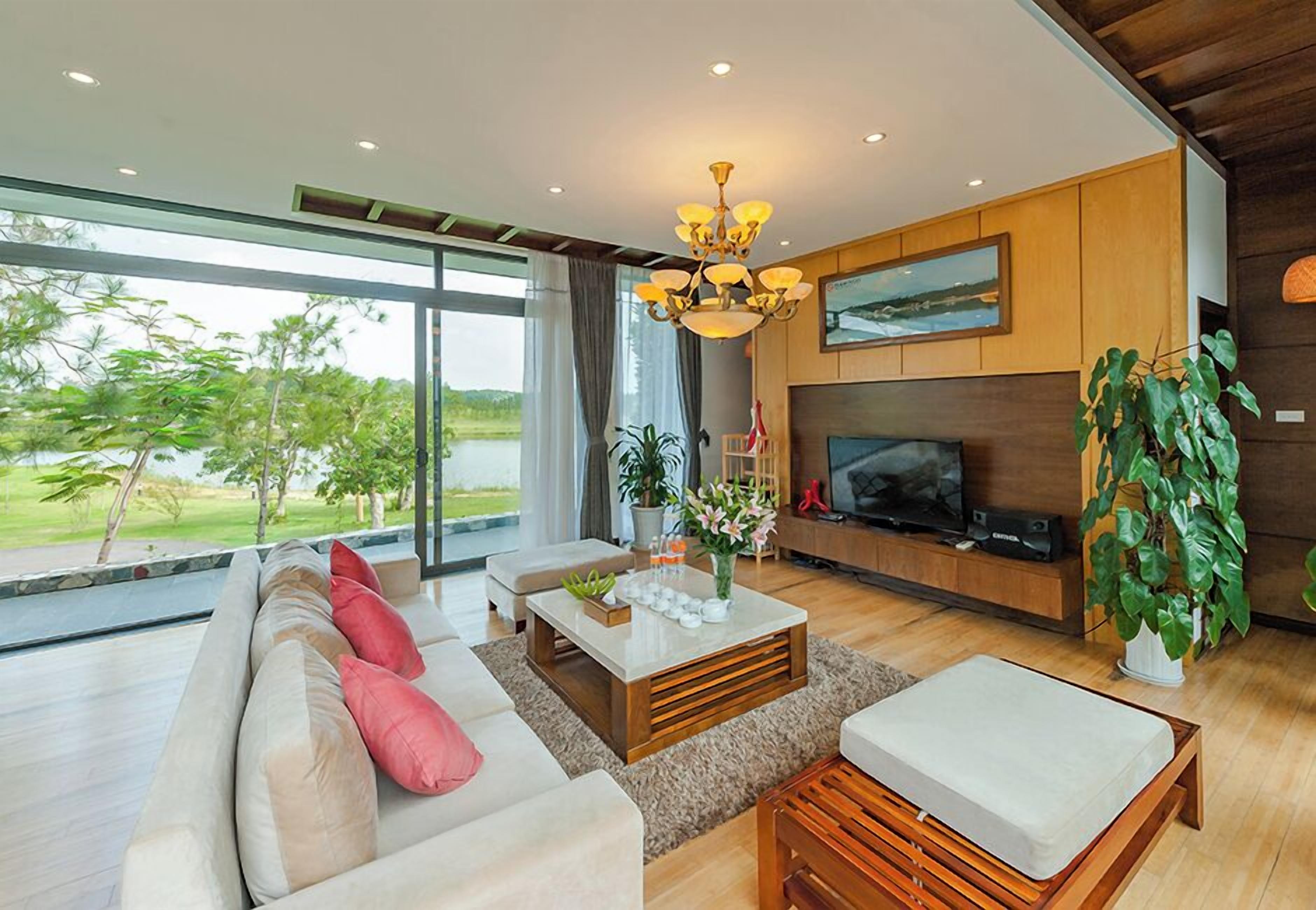 A modern living room with a large sofa, a coffee table, and a TV. There is a large window with a view of trees and a body of water.