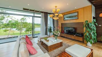 A modern living room with a large sofa, a coffee table, and a TV. There is a large window with a view of trees and a body of water.
