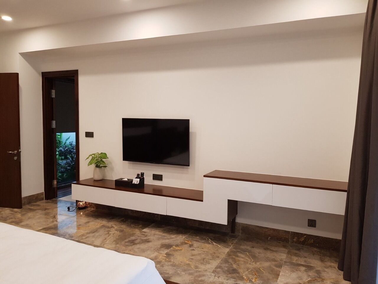 Een moderne slaapkamer met een groot bed, een aan de muur bevestigde televisie, een console met een plant en een deur naar een balkon.