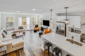 Ein modernes Wohnzimmer mit Sofa, Hocker und Couchtisch, eine KĂŒche mit weiĂen SchrĂ€nken und GerĂ€ten sowie eine groĂe Kochinsel mit Hockern.