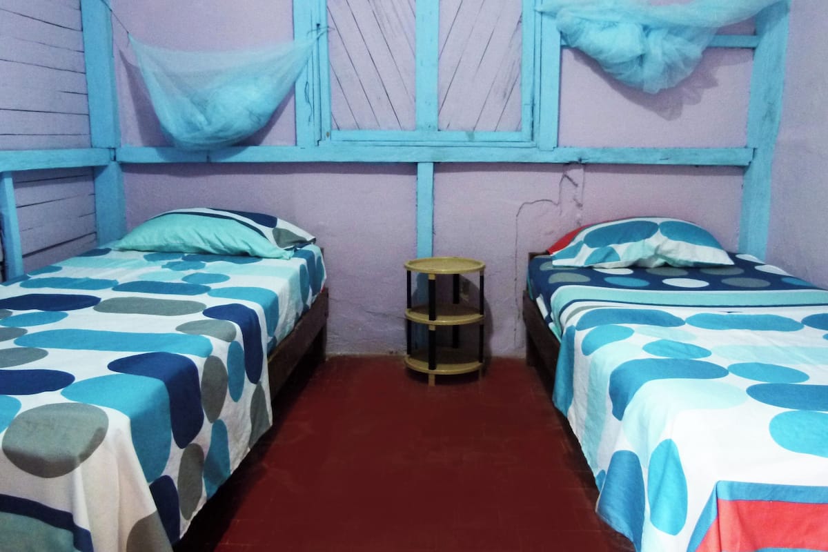 Een kamer met twee bedden, elk met een blauw-wit stippenbedovertrek, een klein rond tafeltje en een raam met een blauw gordijn.