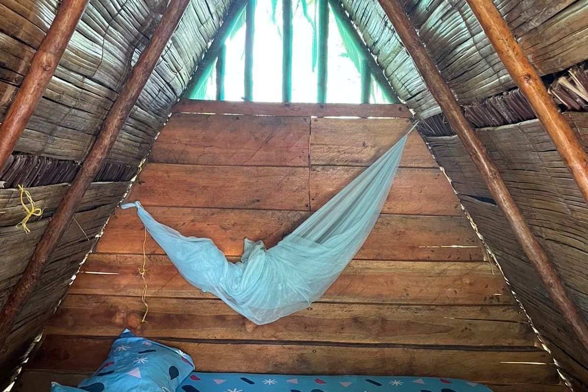 Een hangmat, opgehangen tussen houten balken, in een zolderkamer met rieten dak. Er ligt een matras en kussens met een patroon in blauwtinten op de hangmat.