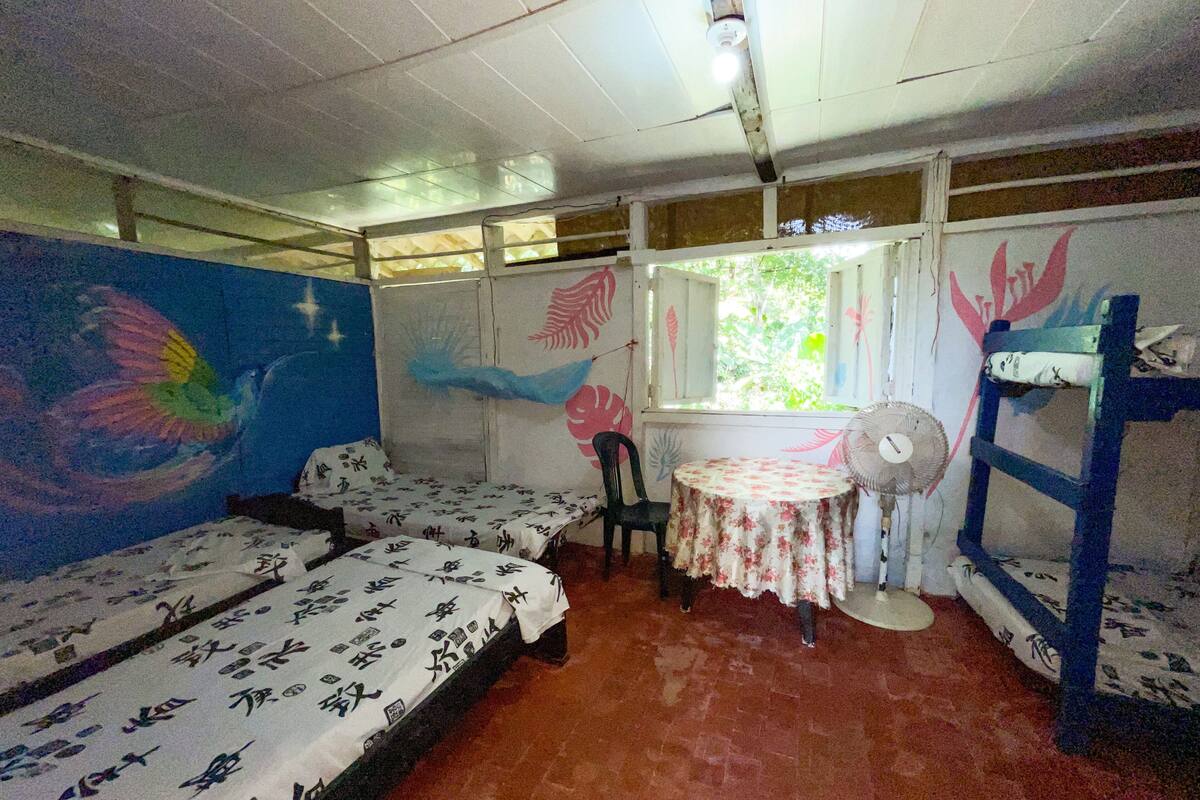 Een kamer met twee bedden, een tafeltje met een ventilator en een raam met uitzicht op groen.