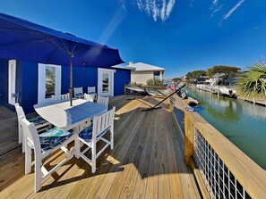 Une terrasse en bois avec une table et des chaises, un auvent bleu et une vue sur une voie navigable où naviguent des bateaux.