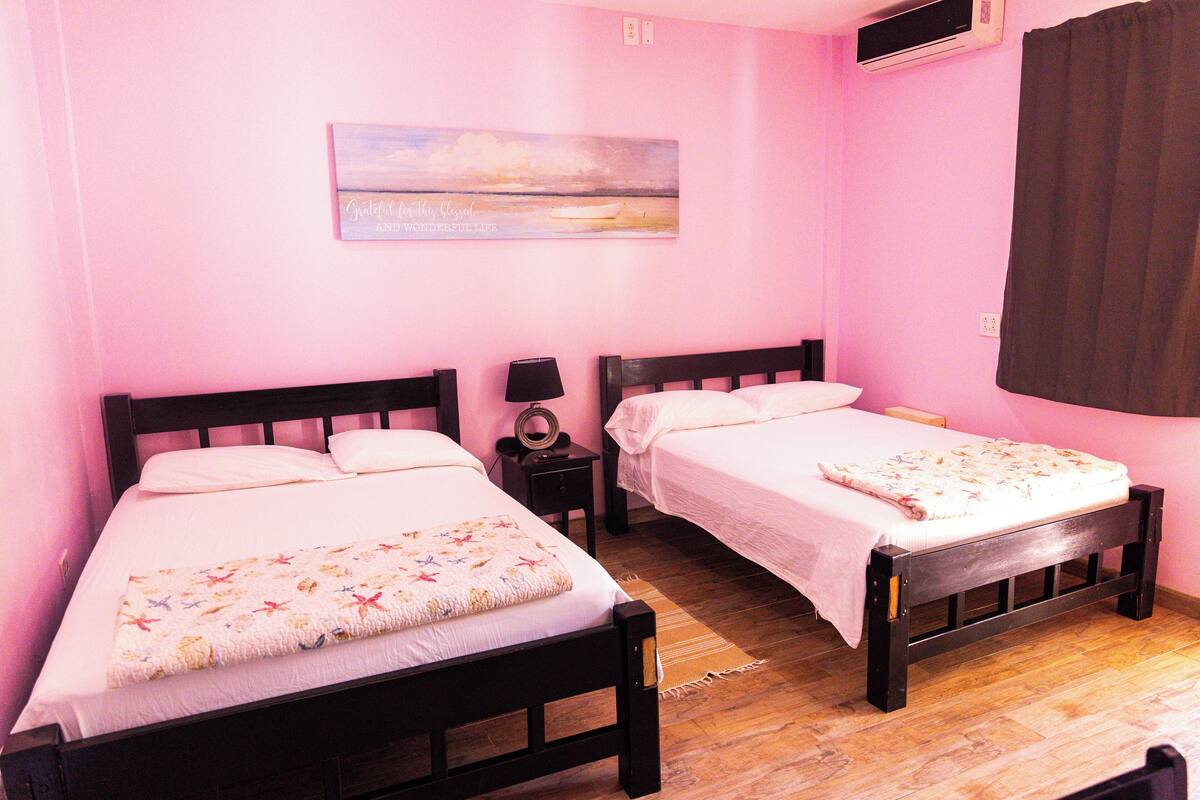 A room with two beds, each with a white bedspread and a floral blanket. There is a painting on the wall above the beds, and an air conditioning unit mounted on the wall.