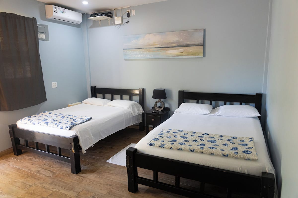 A bedroom with two beds, a wall-mounted air conditioner, a small bedside table with a lamp, and a painting above the beds.