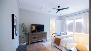 Une chambre à coucher comprenant un lit, un téléviseur, un ventilateur de plafond, une fenêtre avec des rideaux et une porte donnant sur une autre pièce.