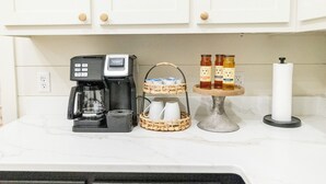 Una encimera de cocina con una cafetera, una cesta con tazas, tarros de miel y un rollo de servilletas de papel.