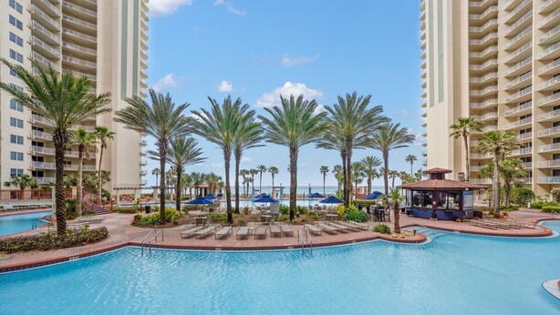 A large swimming pool surrounded by palm trees and lounge chairs, with high-rise buildings in the background. - Beachfront resort condo - pool, hot tub & gym (Panama City Beach)