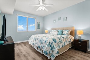A bedroom with a bed, two bedside tables, a ceiling fan, a window with blinds, and a wall with three decorative items. - Beachfront resort condo - pool, hot tub & gym (Panama City Beach)