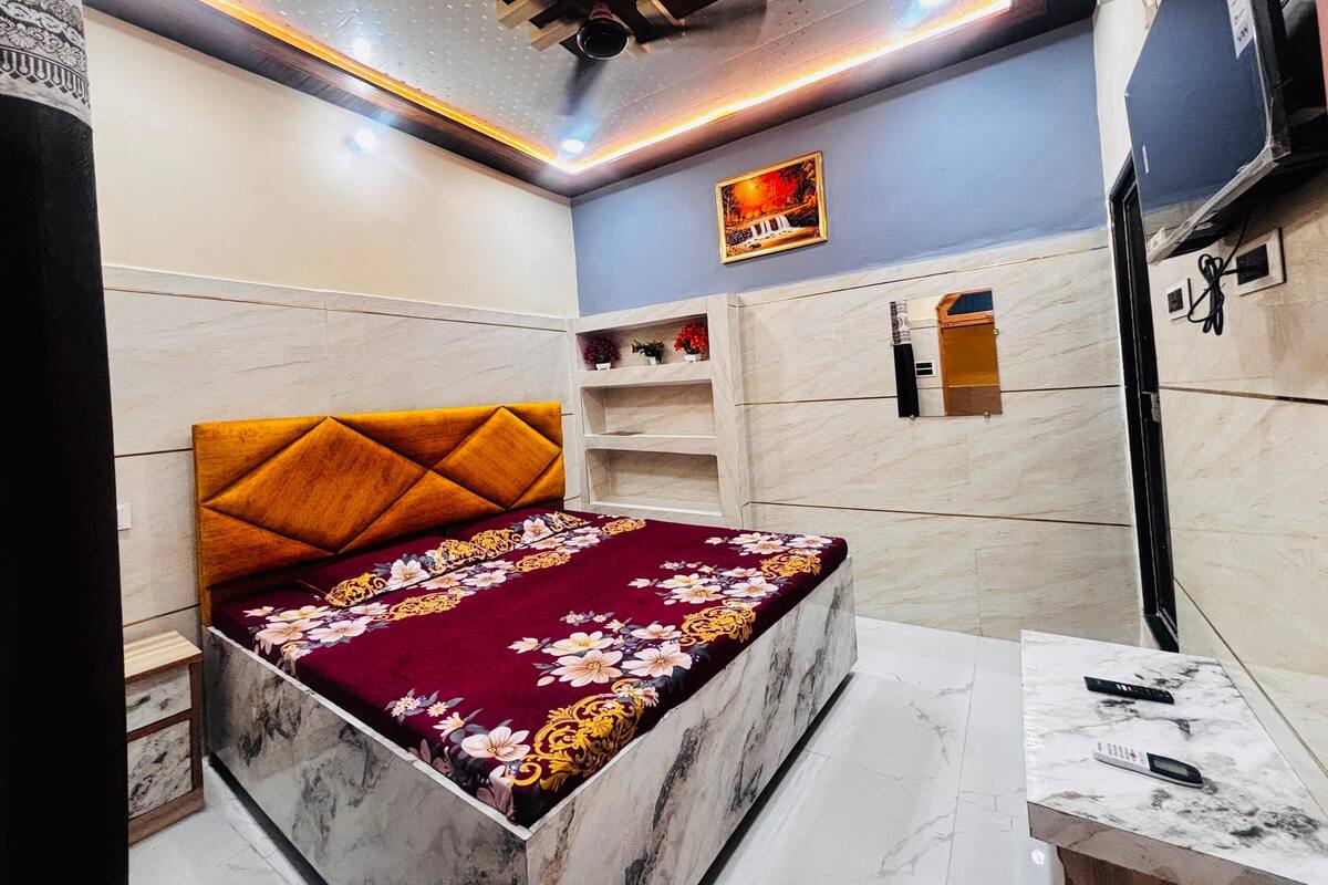 Un dormitorio con cama de mármol, cabecera de madera, colcha con estampado floral, una pequeña estantería con objetos decorativos y un televisor de pared.