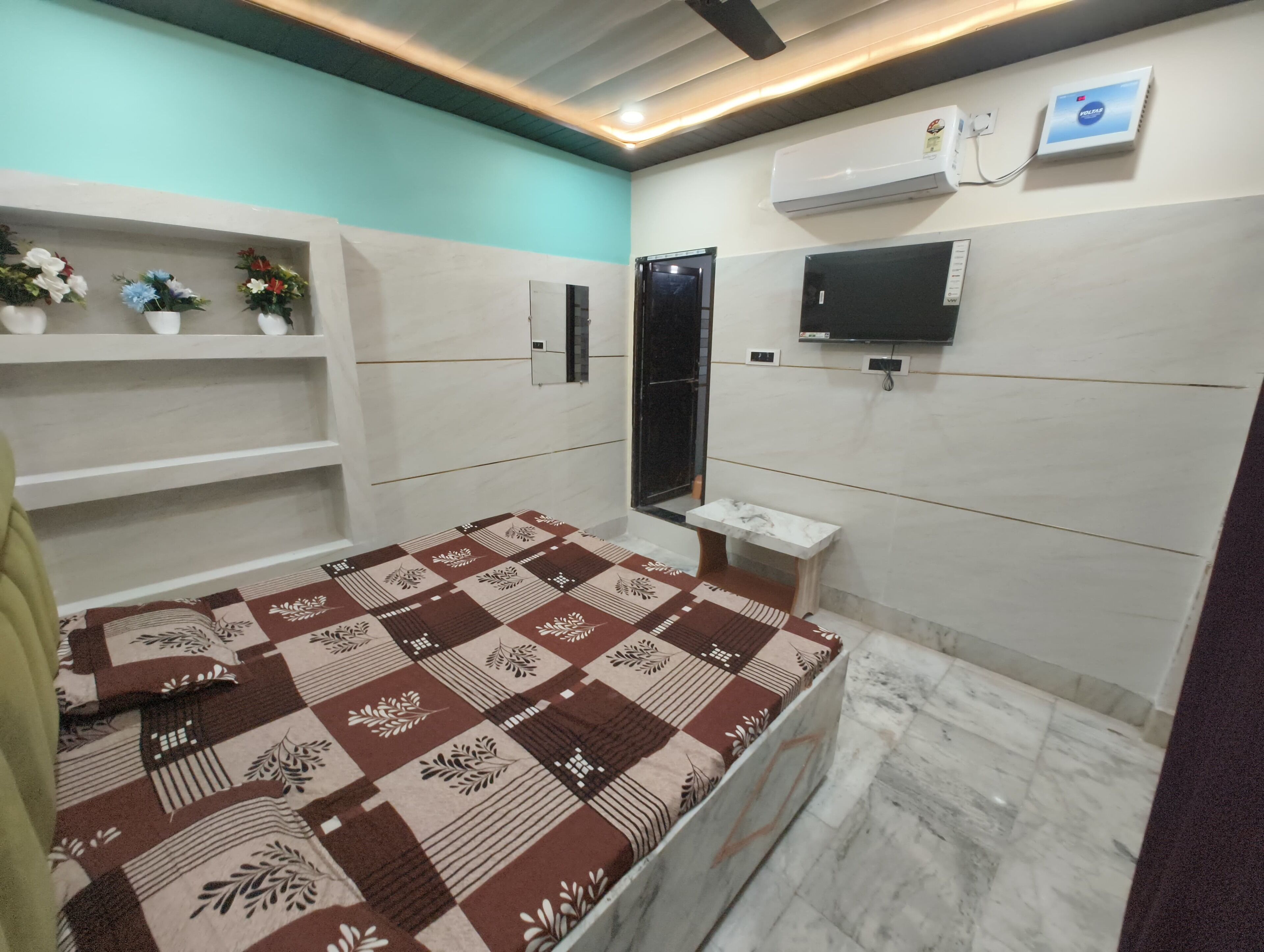 A modern bedroom with a patterned bedspread, a small bedside table, a mounted TV, and an air conditioning unit.