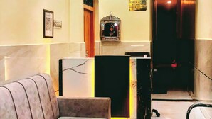 A lobby with a sofa, a reception desk, a ceiling fan, and a framed picture on the wall. - Vatsalya Bhawan Ayodhya (Faizabad)