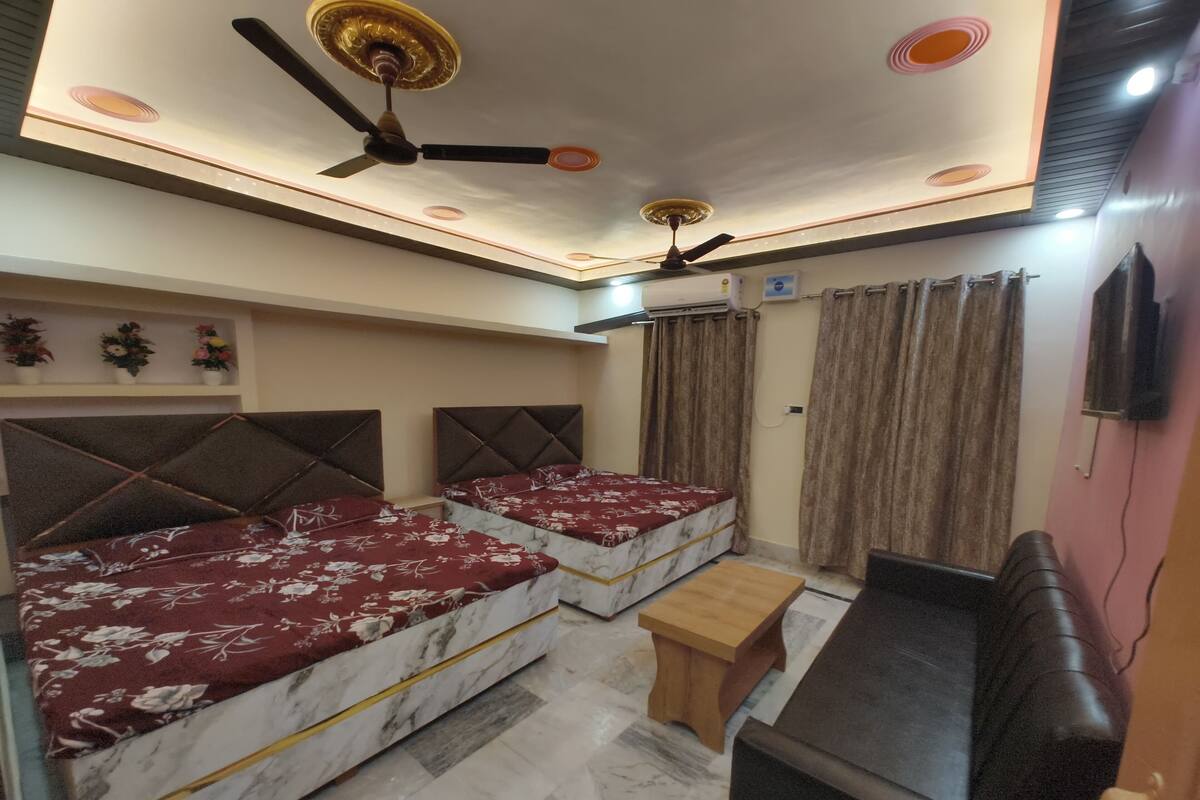Habitación de hotel con dos camas, un banco de madera, ventilador de techo y televisión montada en la pared.