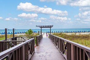 A wooden boardwalk leading to a beach with blue umbrellas and a clear sky.
