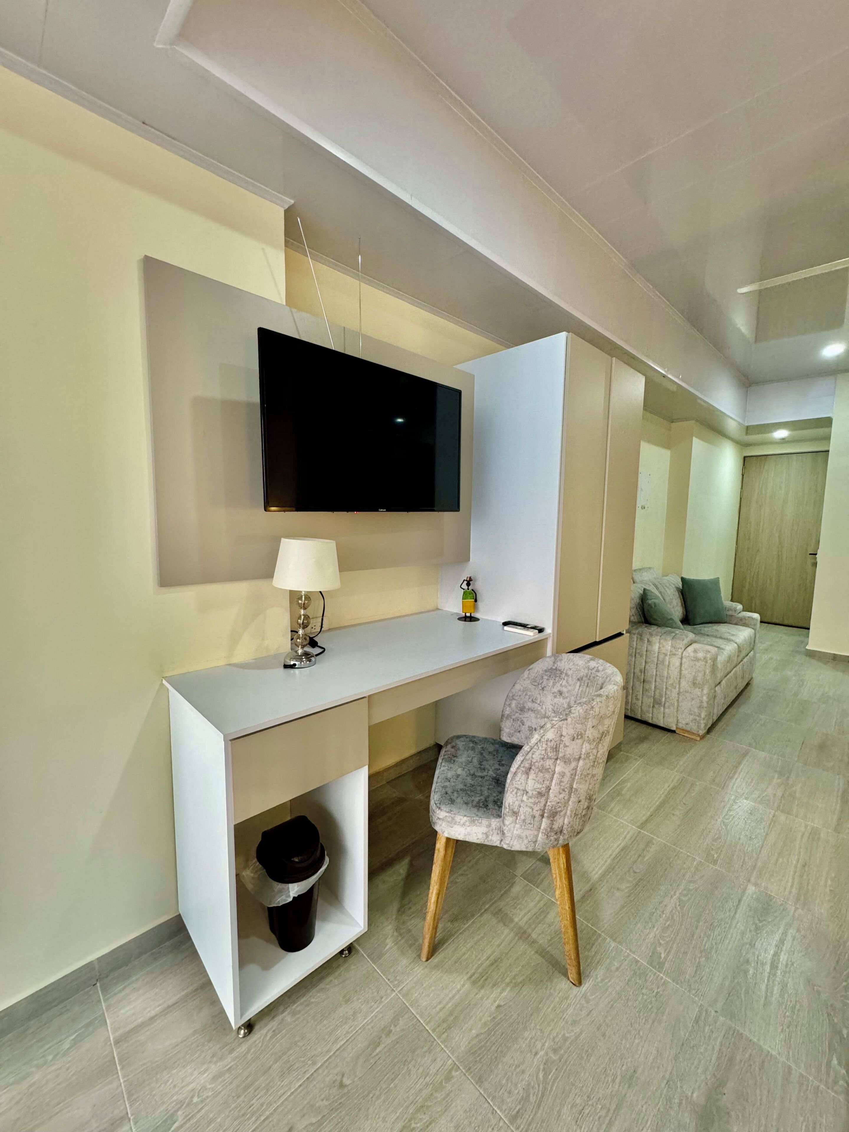 Un salon moderne avec un téléviseur à écran plat fixé au mur, un bureau blanc avec une lampe, un fauteuil gris et un canapé.
