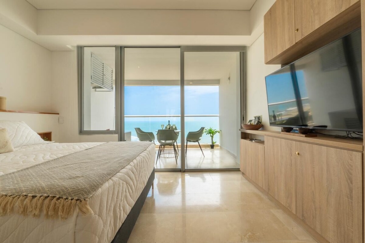 A bedroom with a bed, a TV, a balcony with a table and chairs, and a view of the sea.