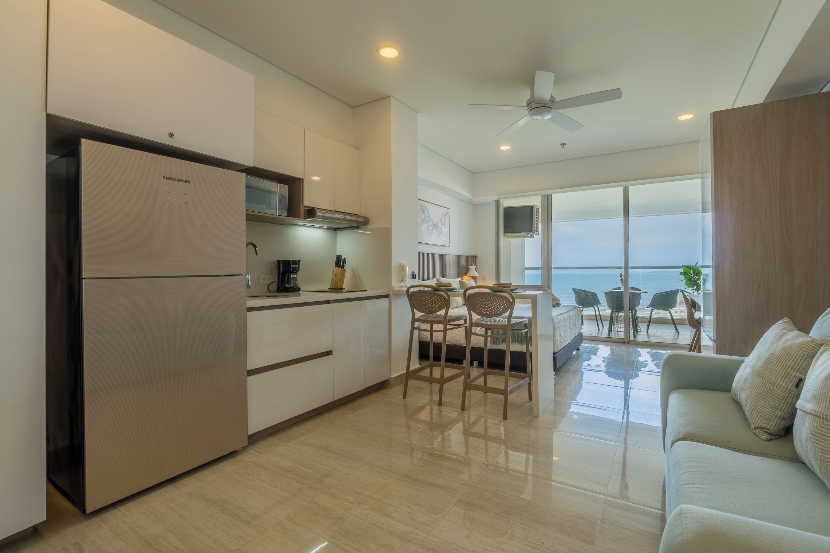 A modern kitchen with a built-in refrigerator, a dining area with chairs, and a view of the sea through large windows.