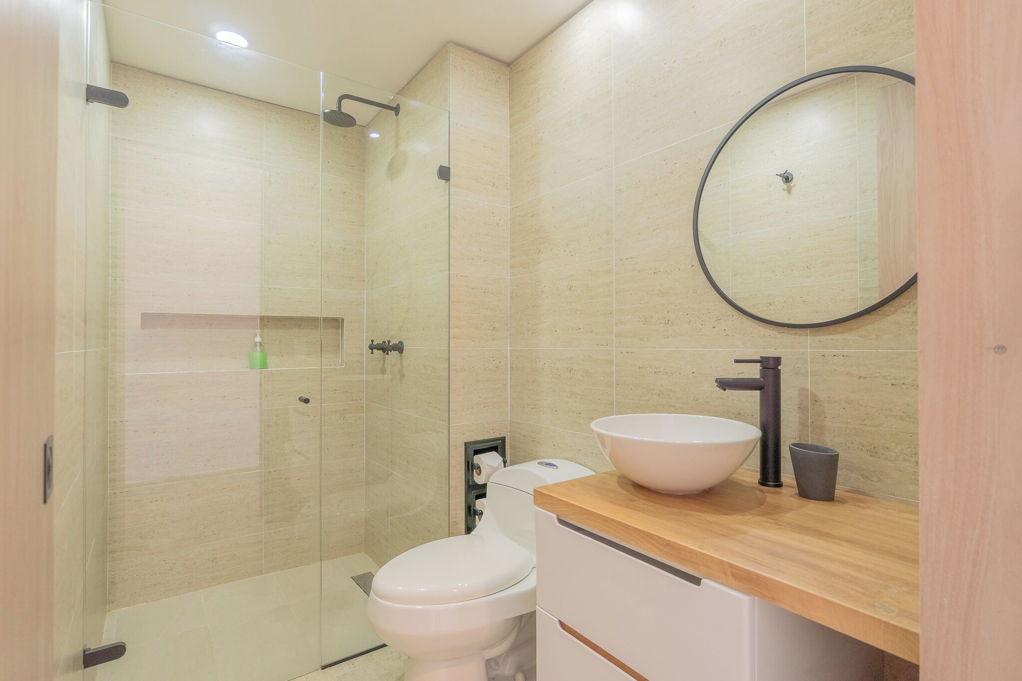 Une salle de bain moderne avec une douche à parois de verre, des toilettes blanches, un meuble-lavabo en bois doté d’un grand évier et un miroir rond.